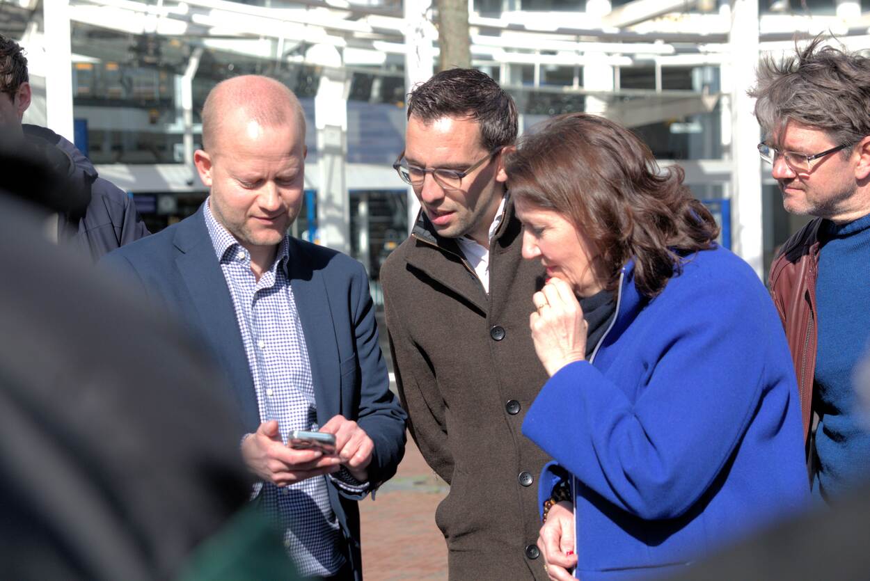 Twee mannen en een vrouw staan bij elkaar en kijken naar beneden. Achter hen staan andere mensen die met elkaar in gesprek zijn.