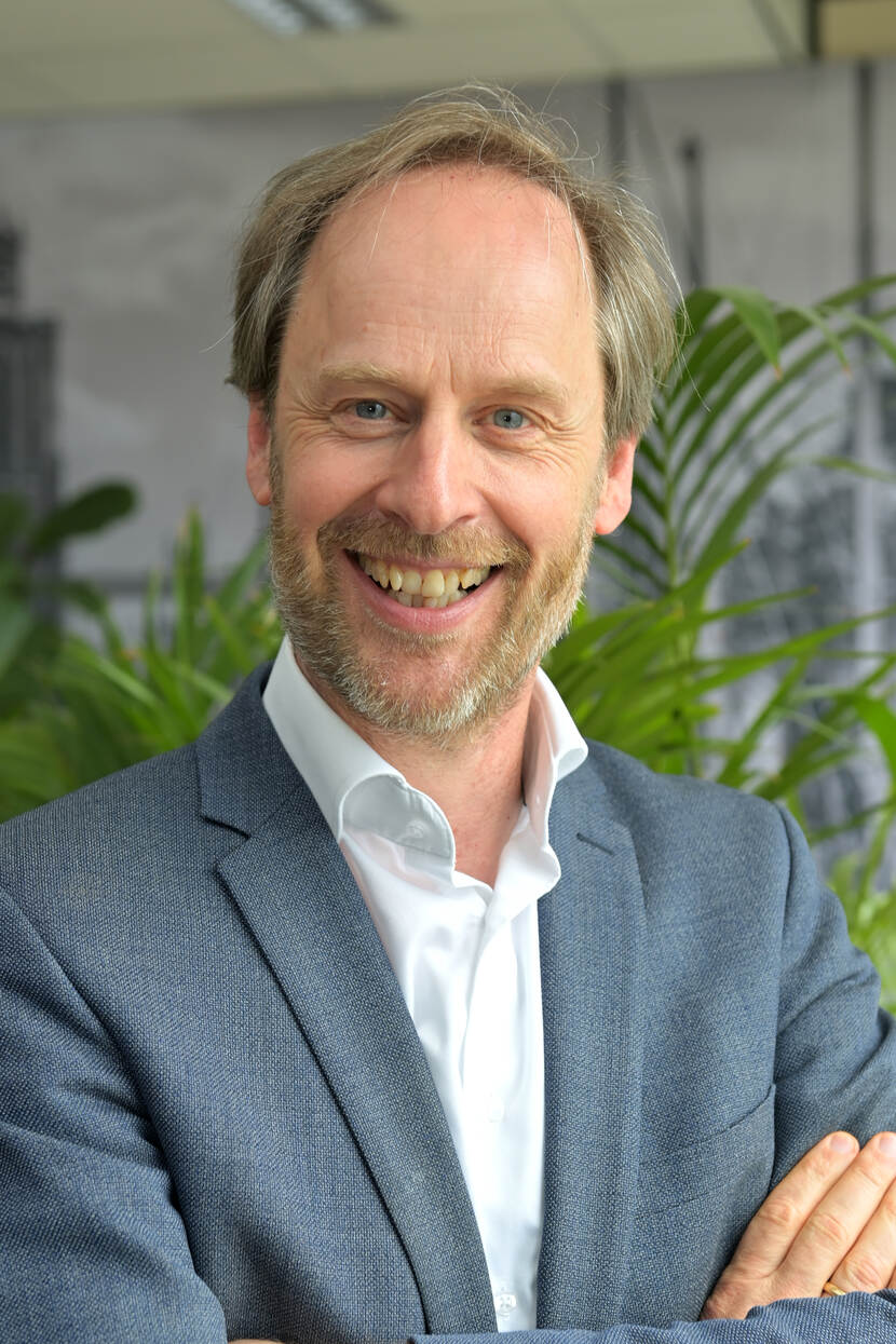 Portretfoto van Hans Goossensen. Hans draagt een grijs colbert met daaronder een witte blouse.